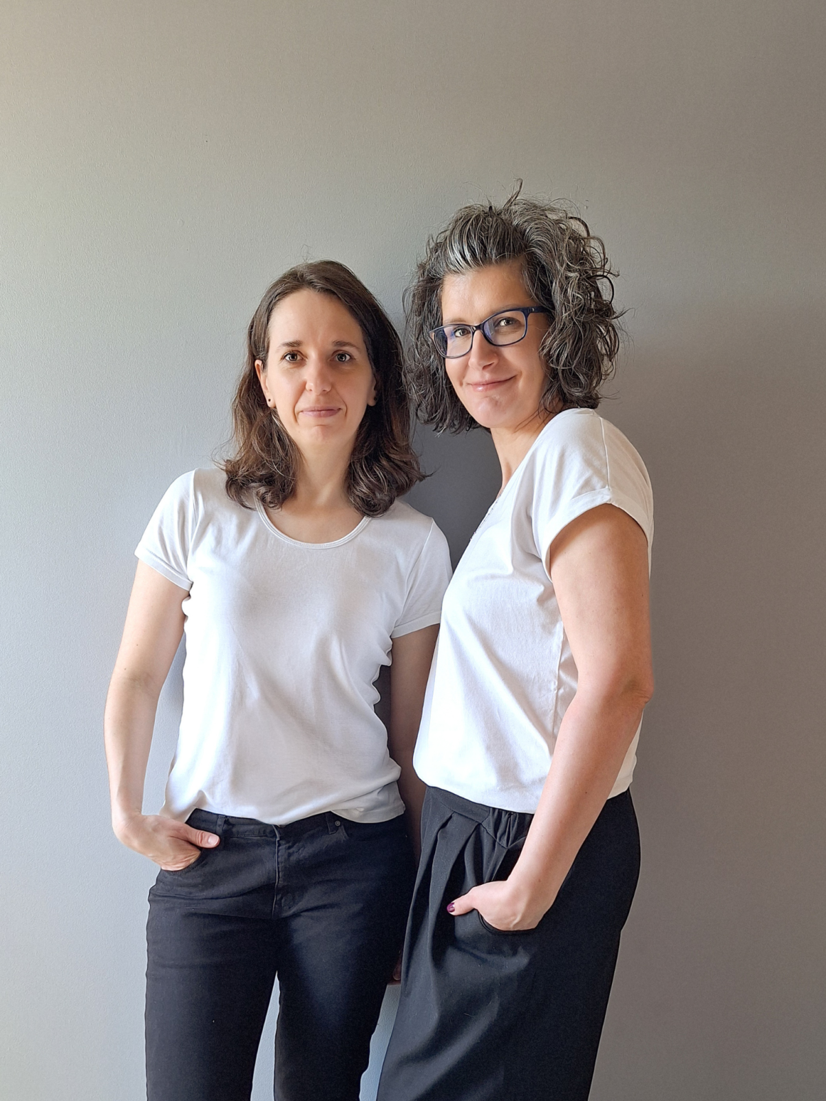 Dwie uśmiechnięte kobiety – zespół Słowo po Słowie, Gosia (w okularach) i Ewelina, stoją obok siebie na szarym tle.