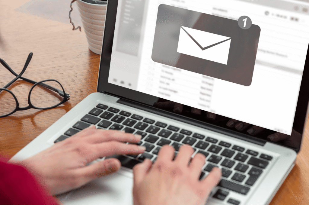 Dłonie piszące na laptopie obok ikony nowej wiadomości e-mail, okulary i kawa na biurku.